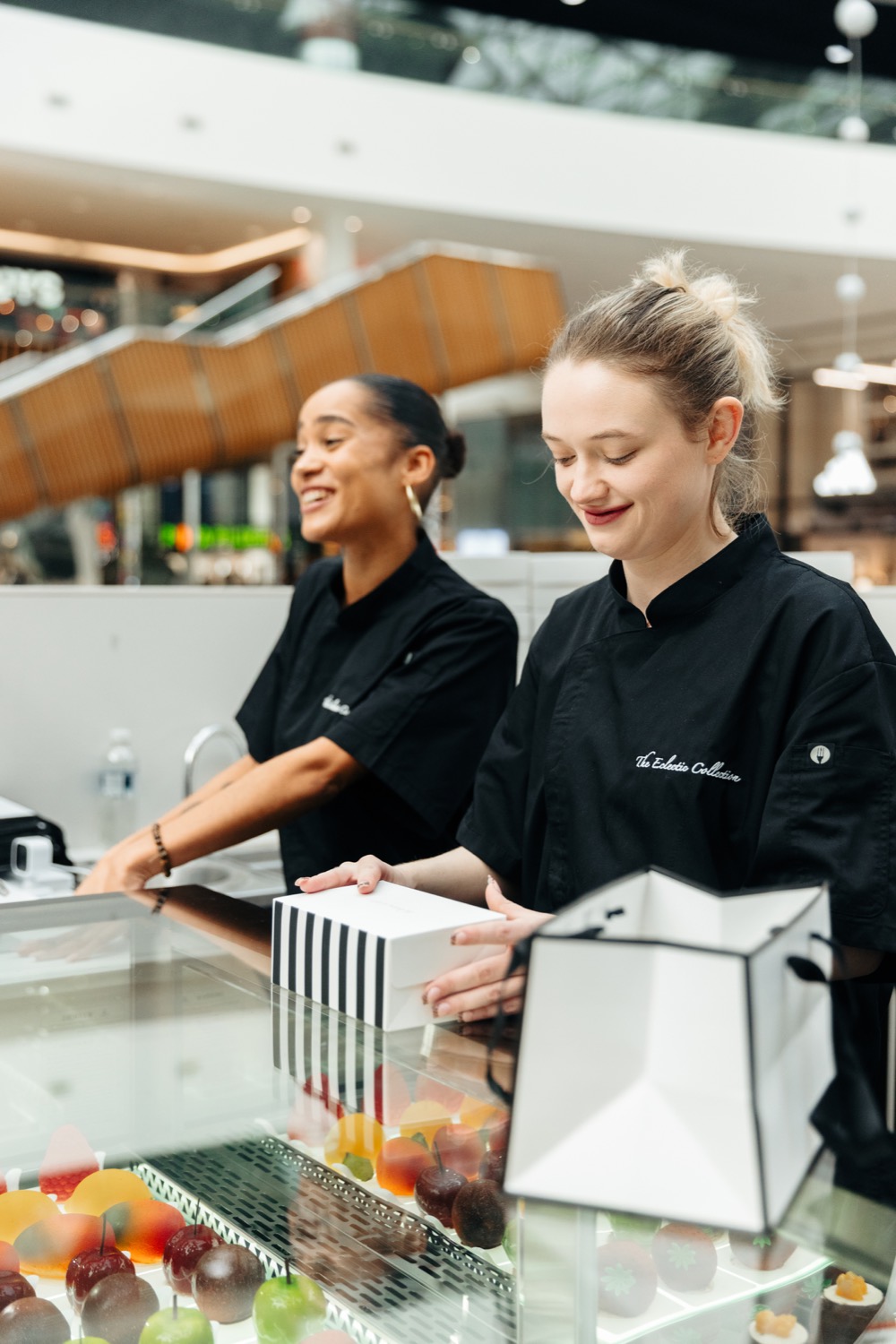 Two members of staff smiling behind the patisserie counter at Westfield, boxing an order over a lit display cabinet