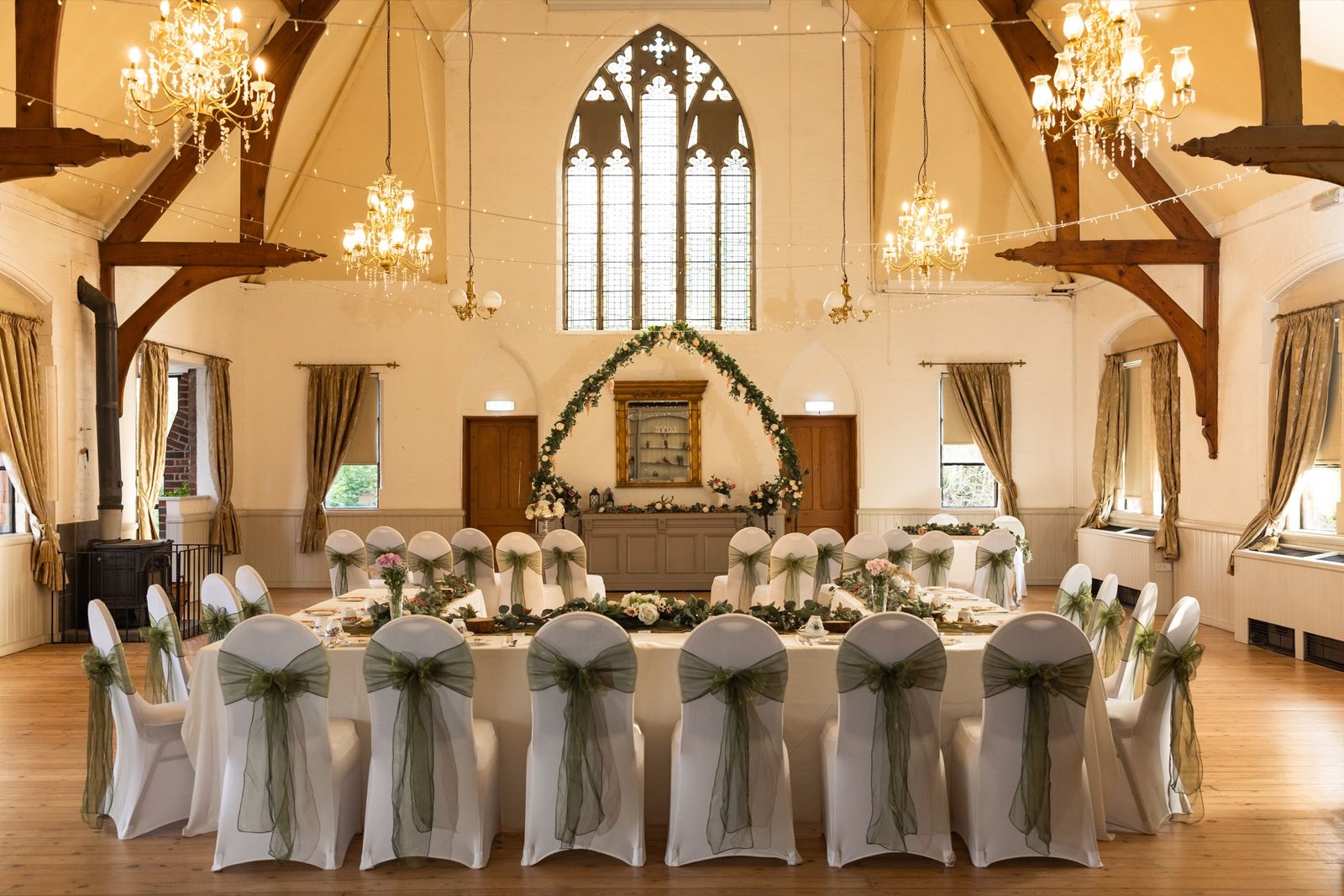 The main hall at Springfield Hall dressed for a wedding breakfast, with exposed timber roof trusses, chandeliers and a Gothic arched window