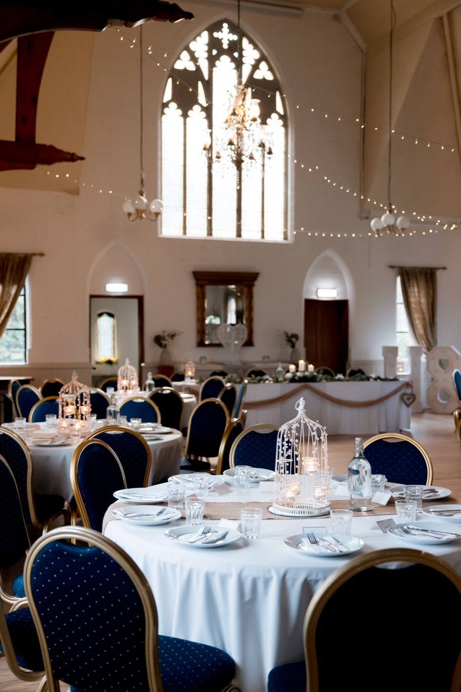 Round tables laid out for a celebration beneath the Gothic window and festoon lighting